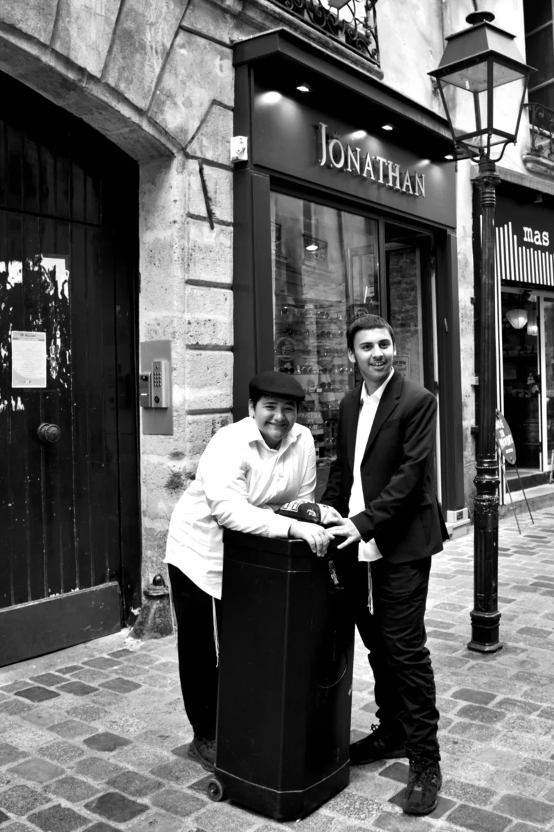 Fabienne · 01/07/2026 · 2925 x 4388 Cette photographie en noir et blanc montre deux personnes posant devant une boutique nommée "Jonathan". Le sujet principal est un couple, l’un tenant un sac, souriants et proches.La composition est symétrique, avec un cadrage serré sur les deux individus. La lumière est naturelle, douce et légèrement contrastée, mettant en valeur les contours.Les tons dominants sont les gris et noirs, typiques du noir et blanc, renforçant une ambiance classique et intemporelle. Le style est sobre et élégant, évoquant une scène urbaine et chaleureuse.