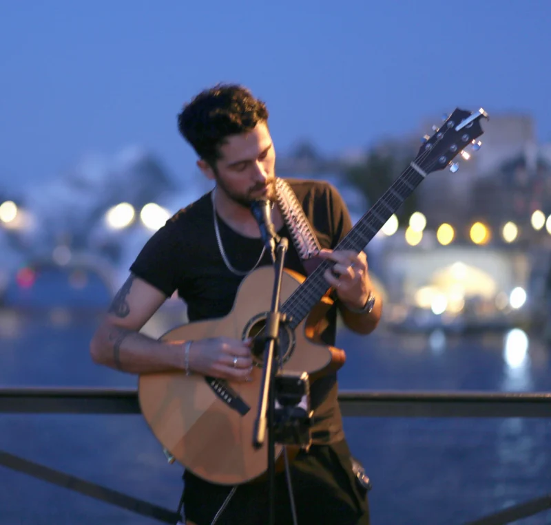 Fabienne · 02/07/2026 · 3828 x 3648 Cette photographie met en scène un homme jouant de la guitare acoustique. Le sujet principal est centré, avec un cadrage serré qui isole son visage et ses mains.La lumière, douce et tamisée, provient probablement des lumières urbaines en arrière-plan, créant une ambiance nocturne et intimiste. Les couleurs dominantes sont des teintes chaudes et bleutées, renforçant l'atmosphère sereine et mélancolique. Le style est naturel et spontané, évoquant une scène de rue ou un moment de performance improvisée. L’ambiance est calme et poétique, presque nostalgique.