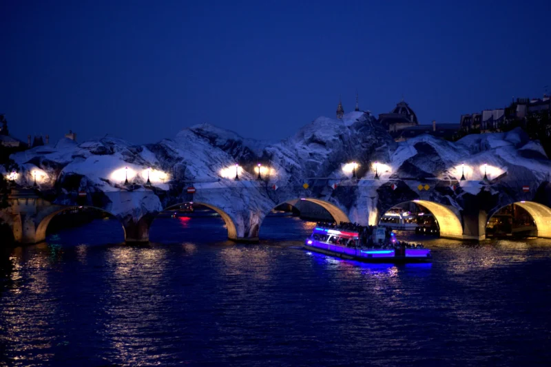 Fabienne · 02/07/2026 · 4631 x 3087 Cette photographie capture un pont illuminé la nuit, dont les arches sont éclairées par des lumières chaudes et bleutées. Le sujet principal est un bateau à moteur dont les phares bleus se reflètent sur l’eau calme.La composition met en valeur le contraste entre les structures architecturales lumineuses et l’obscurité environnante. Le cadrage est serré sur le pont et le bateau, créant une ambiance immersive. Les couleurs dominantes sont les bleus froids et les blancs lumineux, renforçant une atmosphère nocturne et mystérieuse. Le style est réaliste et photographique, évoquant une scène paisible et légèrement magique.