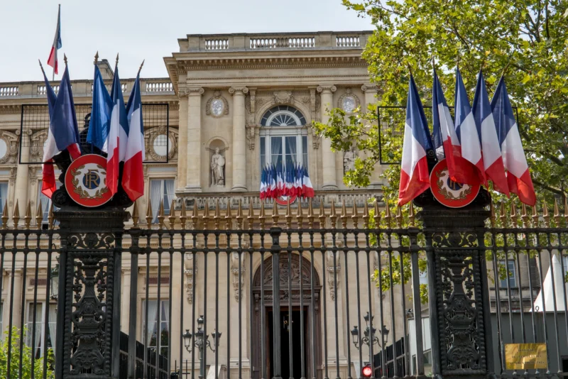 Frédéric · 14/07/2026 · 2517 x 1680 Cette photographie montre l'entrée d'un bâtiment officiel, probablement un ministère français. Le **sujet principal** est l'imposante grille en fer forgé ornée de **drapeaux tricolores** et d'un emblème central.La **composition** est symétrique, avec des drapeaux disposés de chaque côté. Le **cadrage** est serré sur l'entrée, mettant en valeur les détails architecturaux et les couleurs. La **lumière** est naturelle, douce, éclairant les éléments sans ombre marquée. Les **couleurs dominantes** sont le bleu, blanc, rouge des drapeaux, et le noir de la grille.Le **style** est classique et solennel, évoquant une **ambiance** officielle et majestueuse.