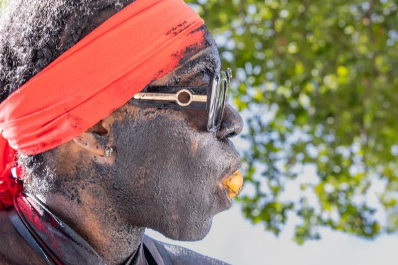 Laurence · 05/07/2026 · 1350 x 900 Cette photographie met en scène un homme comme sujet principal, dont le visage est partiellement recouvert de peinture traditionnelle et de cendres. Il porte un bandeau rouge et des lunettes de soleil.Le cadrage est serré, centré sur son visage, avec un arrière-plan flou de feuillage vert. La lumière est naturelle et semble douce, créant des contrastes subtils. Les couleurs dominantes sont les tons rouges du bandeau, les verts de la végétation et les teintes grises et noires de la peinture corporelle.Le style est documentaire et authentique, évoquant une ambiance culturelle et rituelle.