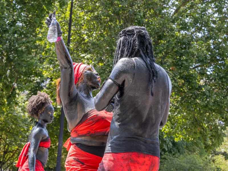 Laurence · 05/07/2026 · 1200 x 900 Cette photographie met en scène trois personnes participant à une cérémonie traditionnelle, probablement aborigène australienne. Le sujet principal est l’homme dos à la caméra, peint de blanc et noir, levant un bras.La composition est dynamique, avec un cadrage serré sur les participants. La lumière naturelle, douce et légèrement filtrée par les arbres, éclaire les visages et les peintures corporelles. Les couleurs dominantes sont les ocres (blanc, noir) et le rouge vif des vêtements.Le style est documentaire et authentique, capturant une ambiance rituelle et culturelle intense, empreinte de spiritualité et de tradition.
