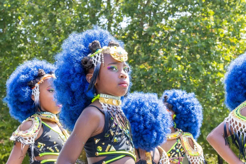 Laurence · 05/07/2026 · 1350 x 900 Cette photographie met en scène un groupe de danseurs traditionnels, dont une jeune fille en premier plan est le sujet principal. Elle porte une coiffure bleue volumineuse et des parures colorées, typiques d’un costume culturel.La composition est dynamique, avec un cadrage serré sur les danseurs en mouvement. La lumière naturelle, douce et légèrement contrastée, éclaire la scène, mettant en valeur les détails des costumes.Les couleurs dominantes sont les bleus vifs des coiffures, les tons dorés et les motifs colorés des vêtements. Le style est traditionnel et festif, créant une ambiance joyeuse et vibrante, évoquant une célébration culturelle.