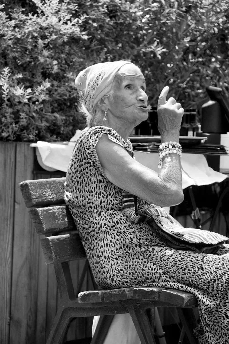 Pascale CHESNOT · 30/05/2026 · 1000 x 1500 Personne âgée assise sur un banc en extérieur, gesticulant avec sa main droite, vêtue d'une tenue à motifs animaux.