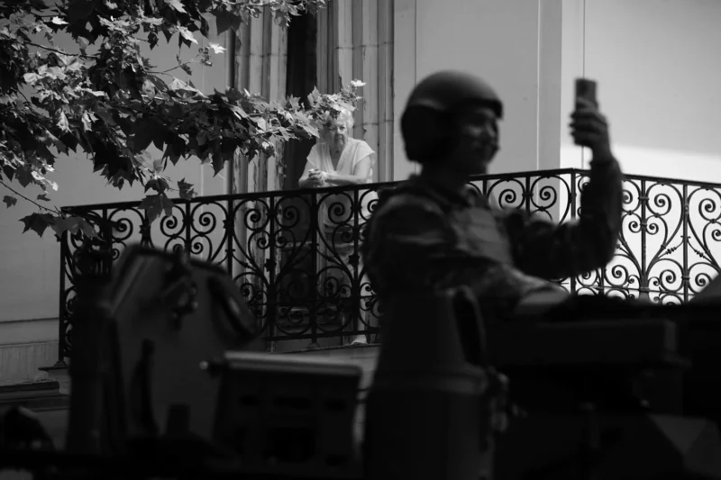 Pascale · 14/07/2026 · 1500 x 1000 Personne utilisant un téléphone devant une balustrade en métal avec une autre personne assise en arrière-plan sous un arbre.