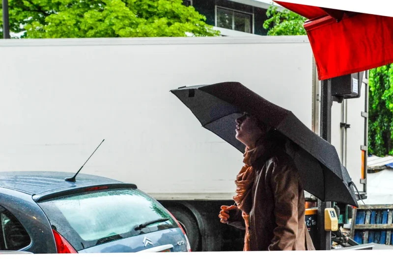 Léonard · 1023 x 685 Personne tenant un parapluie noir sous une bâche rouge près d'une station-service, avec une voiture en arrière-plan sous