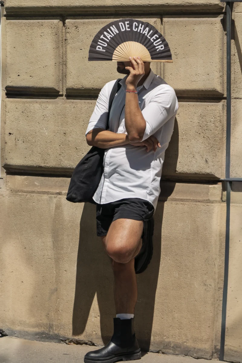 Thierry · 06/07/2026 · 2560 x 3840 Personne utilisant un éventail personnalisé avec écriture en français devant un mur en pierre, vêtue d'un short et d'une