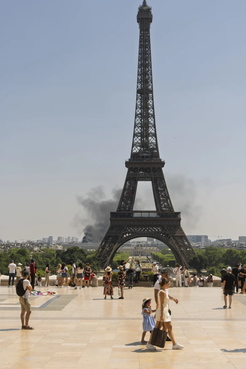 Thierry · 08/07/2026 · 2560 x 3840 Tour Eiffel avec une épaisse fumée noire s'élevant derrière elle, vue depuis son pied avec des visiteurs sur une place