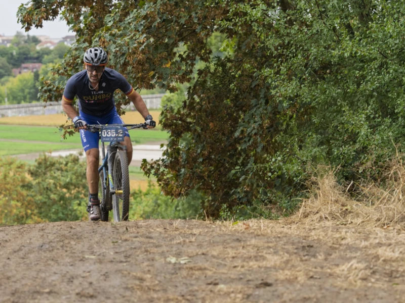 Cette photographie montre un cycliste en mouvement sur un sentier terreux, vêtu d’une tenue sportive et équipé d’un casque. Le sujet principal est net et centré, avec un cadrage dynamique suggérant l’action.La composition utilise un premier plan légèrement flou pour mettre en valeur le cycliste. La lumière naturelle, douce et légèrement latérale, éclaire la scène sans créer d’ombres marquées. Les couleurs dominantes sont les verts naturels de la végétation et les tons neutres de la terre et du vélo.L’ambiance est sportive et sereine, évoquant une balade ou une randonnée en pleine nature.