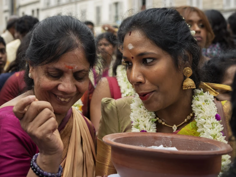 Thierry · 30/08/2026 · 3840 x 2560 Deux femmes souriantes participant à une cérémonie religieuse avec des fleurs et un récipient, entourées d'une foule