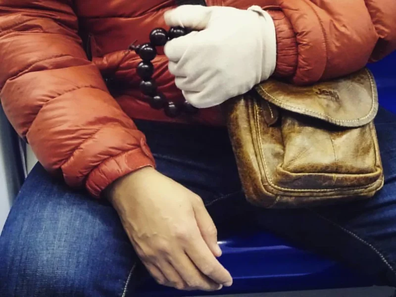 Léonard · 1024 x 768 Personne vêtue d'un manteau orange et de gants blancs tenant un rosaire noir, assise près d'un sac en cuir sur un siège bleu