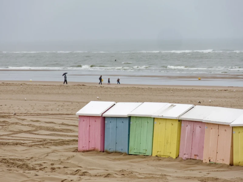 Yvon · 19/08/2026 · 4000 x 2250 Six cabines de plage colorées alignées sur une étendue de sable sous un ciel couvert, avec des vagues à l’arrière-plan