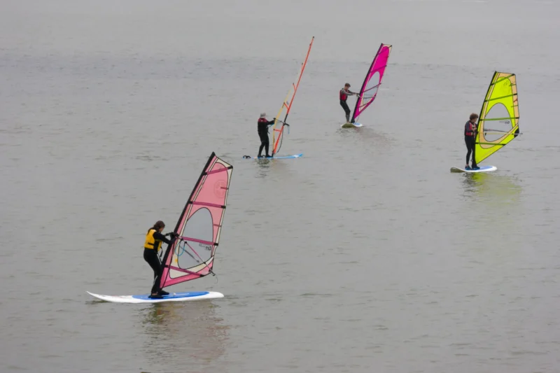 Yvon · 19/08/2026 · 3999 x 2666 Quatre personnes pratiquant le windsurf sur une étendue d'eau calme avec des voiles colorées sous un ciel couvert