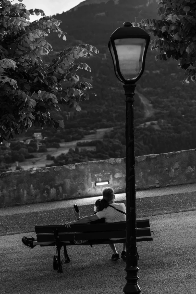 Marc L. · 12/07/2026 · 3393 x 5090 Deux personnes assises sur un banc près d'un lampadaire, observant un paysage avec des arbres et une montagne