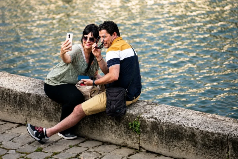 Bao · 20/08/2026 · 1536 x 1024 Deux personnes assises sur une margelle de pierre près de l'eau, prenant un selfie avec un smartphone.