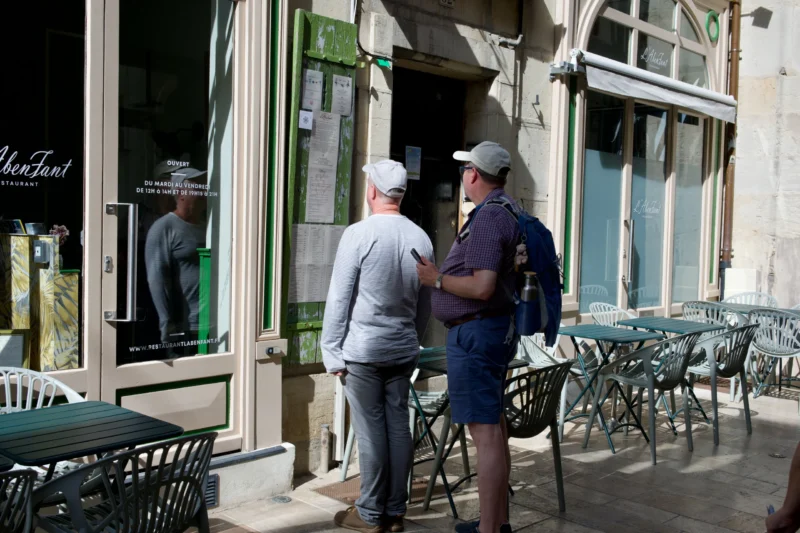 Jean Claude · 24/07/2026 · 6048 x 4032 Deux hommes discutent devant l'entrée d'un restaurant avec des menus affichés à la porte, tables et chaises disposées