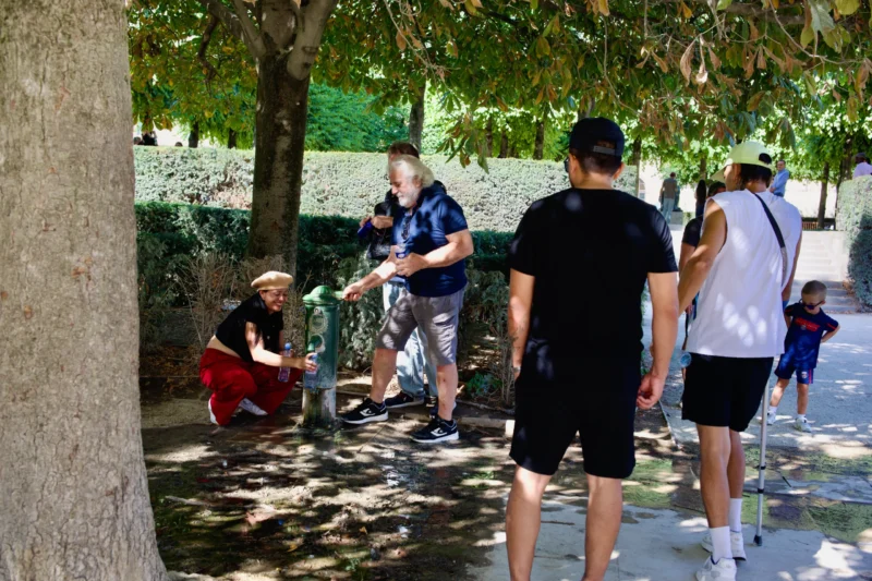 Jean Claude · 09/08/2026 · 6048 x 4032 Quatre personnes près d’un point d’eau dans un parc arboré, dont une s’accroupit et une autre utilise un téléphone portable.