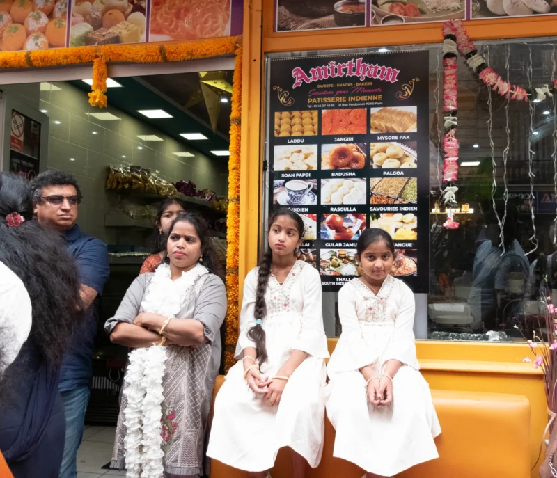 Léonard · 30/08/2026 · 3494 x 3000 Quatre personnes posant devant une vitrine de pâtisserie indienne décorée de guirlandes, avec des produits alimentaires