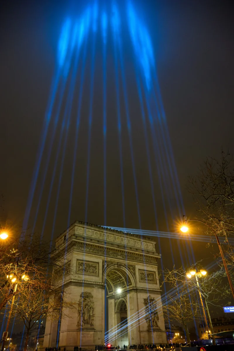 Daniel · 3226 x 4838 Arc de Triomphe illuminé par des faisceaux lumineux bleus traversant la nuit, avec des lampadaires éclairant les arbres
