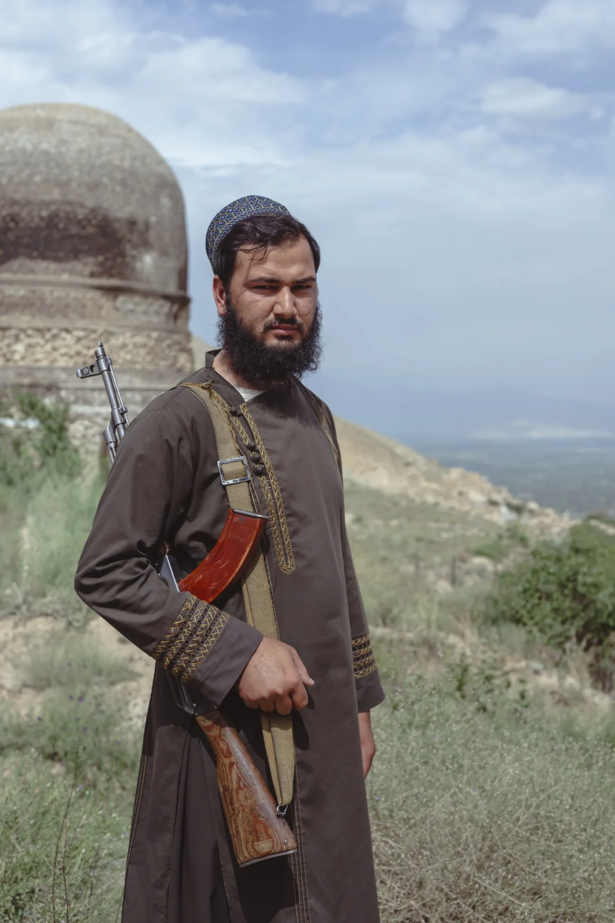 Daniel · 26/05/2026 · 2560 x 3840 Un homme en tenue traditionnelle tenant une arme légère devant un paysage montagneux et des ruines anciennes.