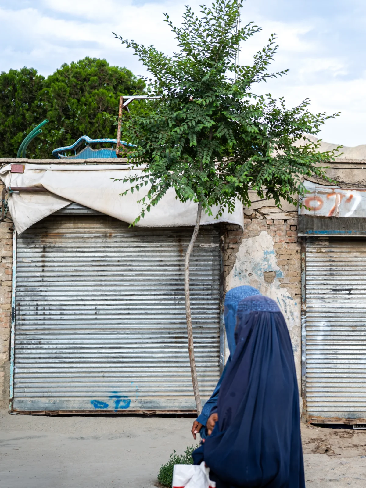 Daniel · 27/05/2026 · 3024 x 4032 Une personne vêtue d'une longue robe bleue marchant devant des bâtiments fermés avec des stores métalliques, sous un arbre