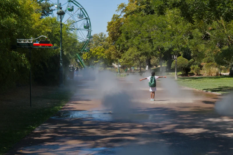 Frédéric · 07/08/2026 · 2518 x 1680 Un enfant court en écartant les bras sous un jet de brume dans une allée ombragée d’un parc avec une roue ferris