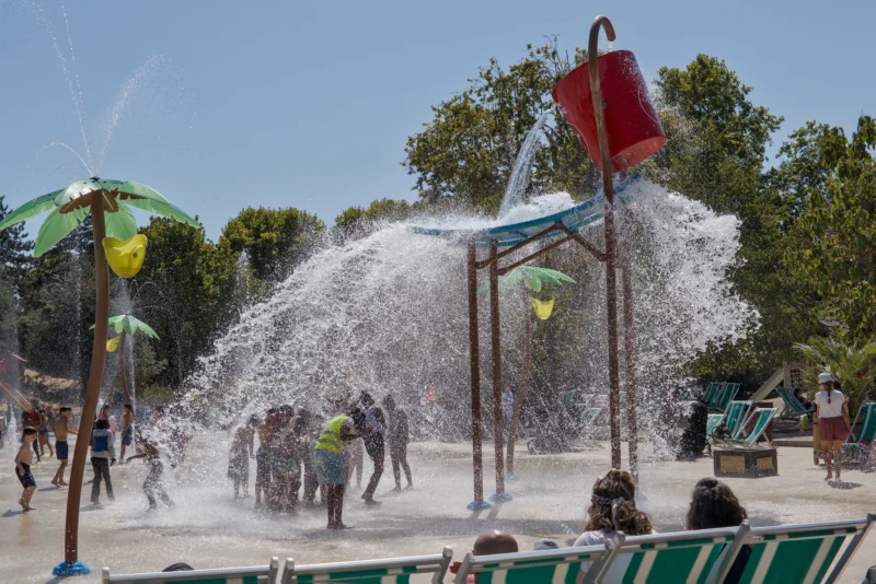 Frédéric · 07/08/2026 · 2517 x 1680 Un groupe de personnes s'amuse dans une zone de jeux d'eau en plein air avec des structures colorées et des jets d'eau