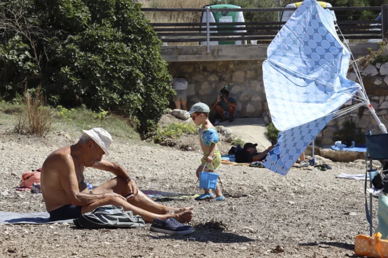 Camilla · 30/08/2026 · 3840 x 2560 Un homme assis sur une serviette au bord d’un chemin de terre près d’un parasol, tandis qu’un enfant joue avec un seau