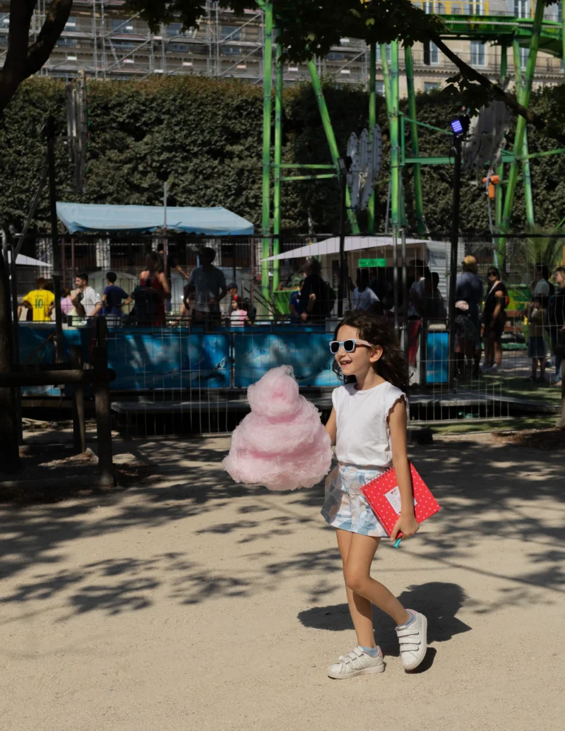 Muriel · 25/07/2026 · 1583 x 2048 Enfant souriante en tenue estivale tenant une grosse barbe à papa rose dans un parc d'attractions ensoleillé
