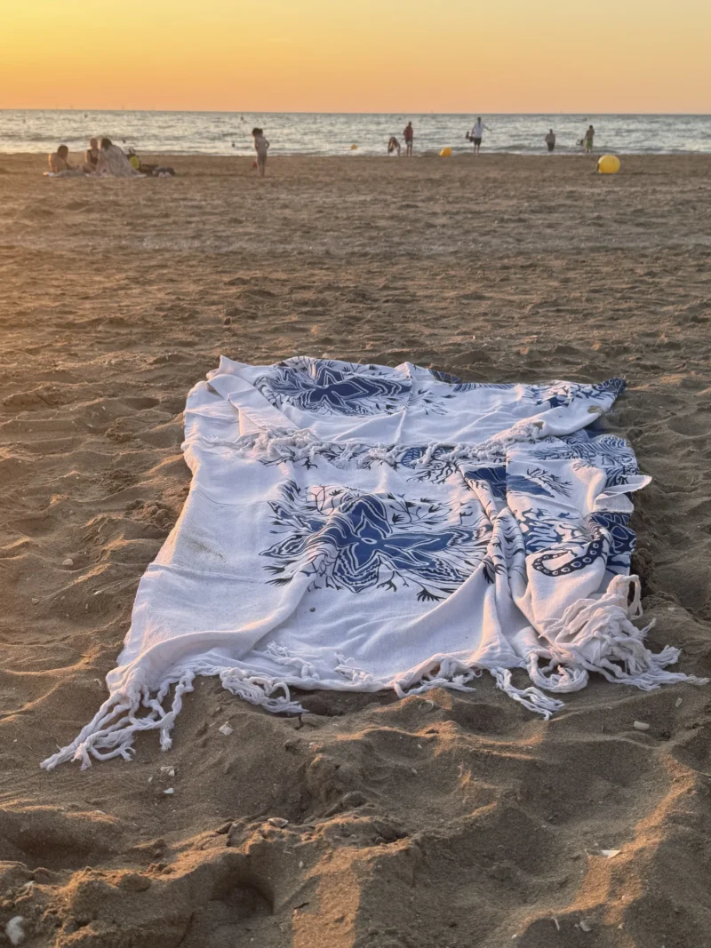 Heike · 12/08/2026 · 1500 x 2000 Serviette de plage à motifs bleus et blancs étendue sur le sable près de l'eau, avec des silhouettes de personnes