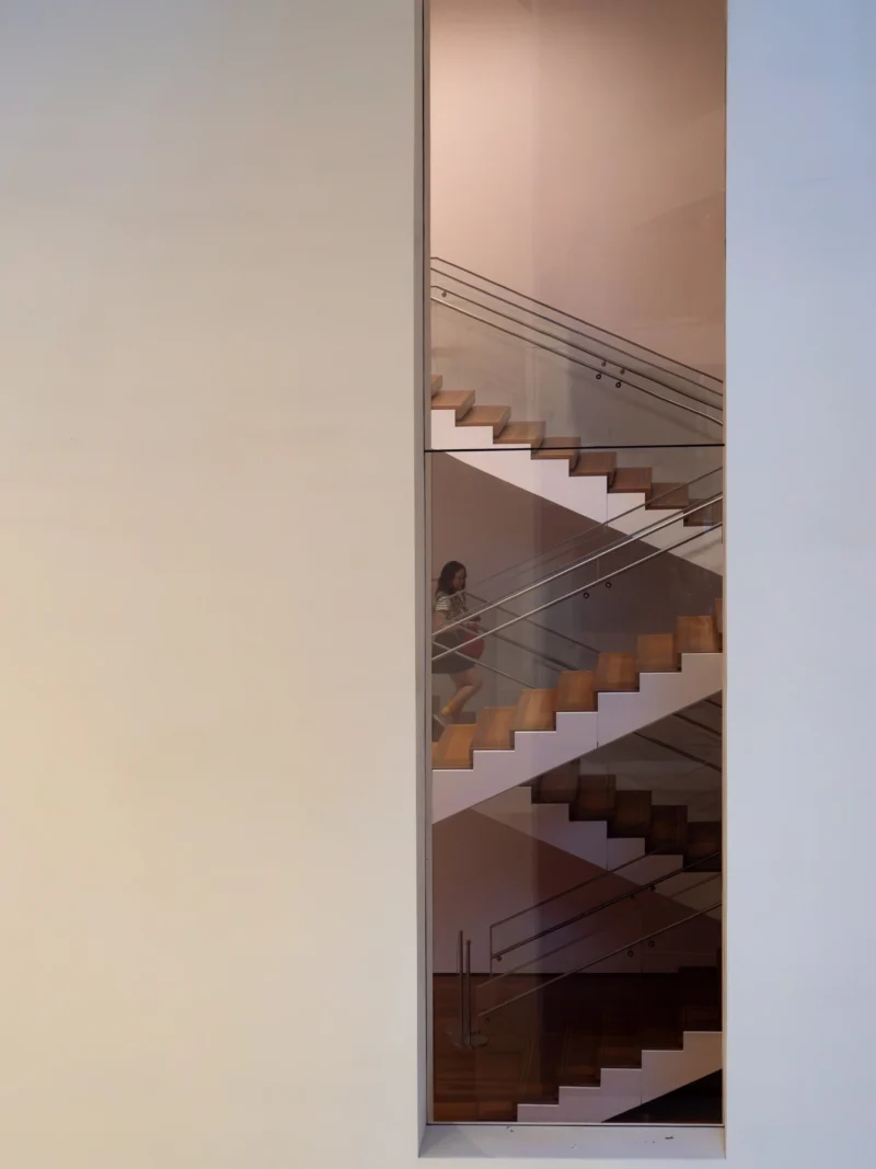 Daniel · 3148 x 4197 Reflet d'un escalier moderne en verre et bois avec une personne assise sur une marche, vue à travers une surface vitrée