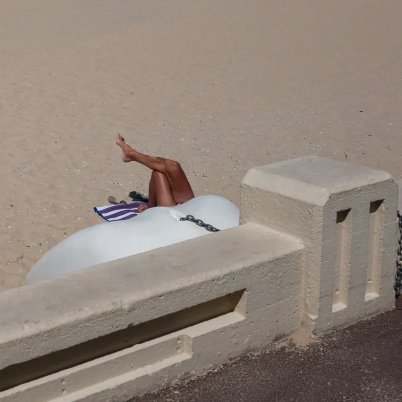 Jean-Philippe · 21/07/2026 · 3840 x 3840 Personne allongée sur un ballon blanc près d’un muret en béton sur une plage, pieds en l’air, avec un tissu coloré à côté.
