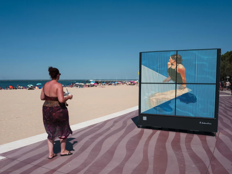 Jean-Philippe · 21/07/2026 · 3840 x 2560 Une personne en tenue légère observe une projection géante sur une structure urbaine près d’une plage ensoleillée avec