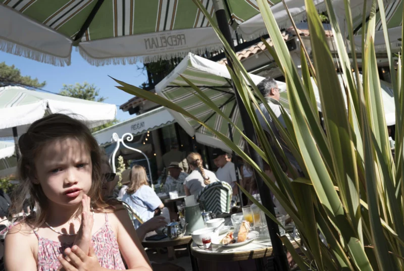 Jean-Philippe · 22/07/2026 · 3840 x 2576 Enfant devant un restaurant extérieur, tenant un objet, avec des tables couvertes et des plantes en premier plan