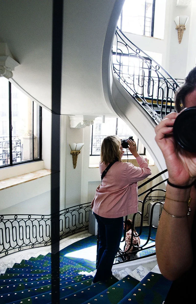 Deux personnes sur un escalier en colimaçon photographiant une vue à travers des fenêtres, dans un bâtiment élégant.