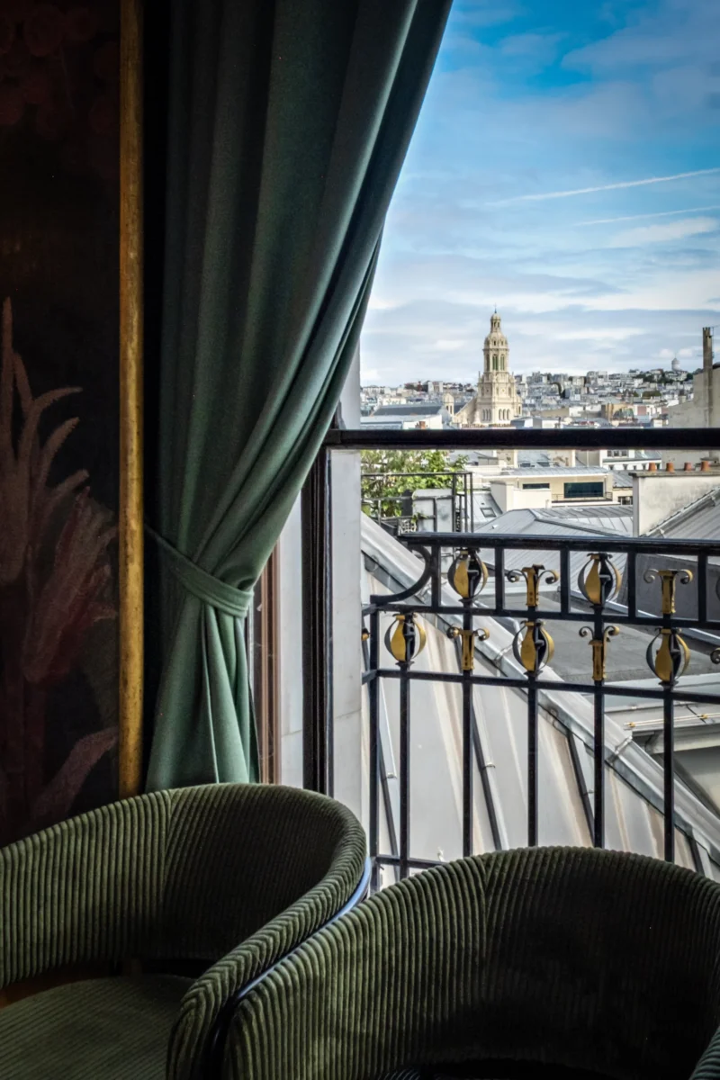 Vue depuis un balcon parisien avec fauteuils verts, balustrade dorée et cathédrale en arrière-plan sous un ciel dégagé.