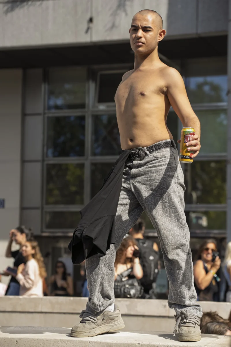 Thierry · 19/09/2026 · 2560 x 3840 Un homme torse nu, vêtu de pantalons larges et de baskets, tient une canette en posant sur une scène devant un public
