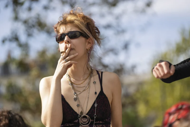 Thierry · 19/09/2026 · 3840 x 2560 Une personne en tenue estivale fume une cigarette en extérieur, avec des accessoires colorés et des lunettes de soleil