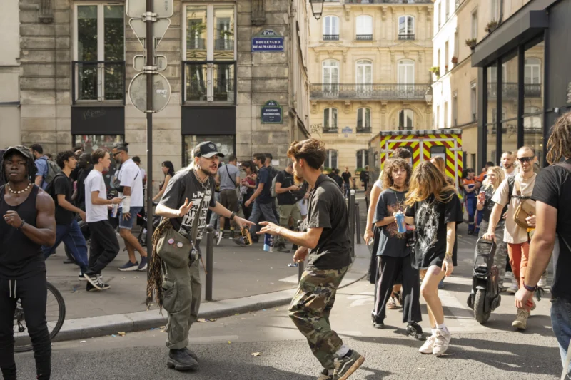 Thierry · 19/09/2026 · 3840 x 2560 Scène animée de rue avec des personnes en mouvement, dont un individu en train de faire du skateboard, dans un quartier