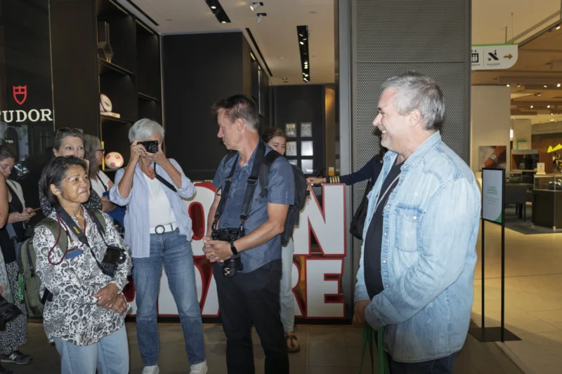 Groupe de personnes en discussion devant une vitrine de magasin avec un panneau lumineux rouge et blanc, ambiance urbaine
