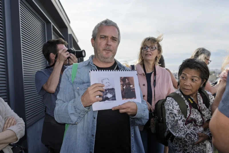 Un homme tient une feuille avec des images devant un groupe de personnes en extérieur, participant à une visite guidée