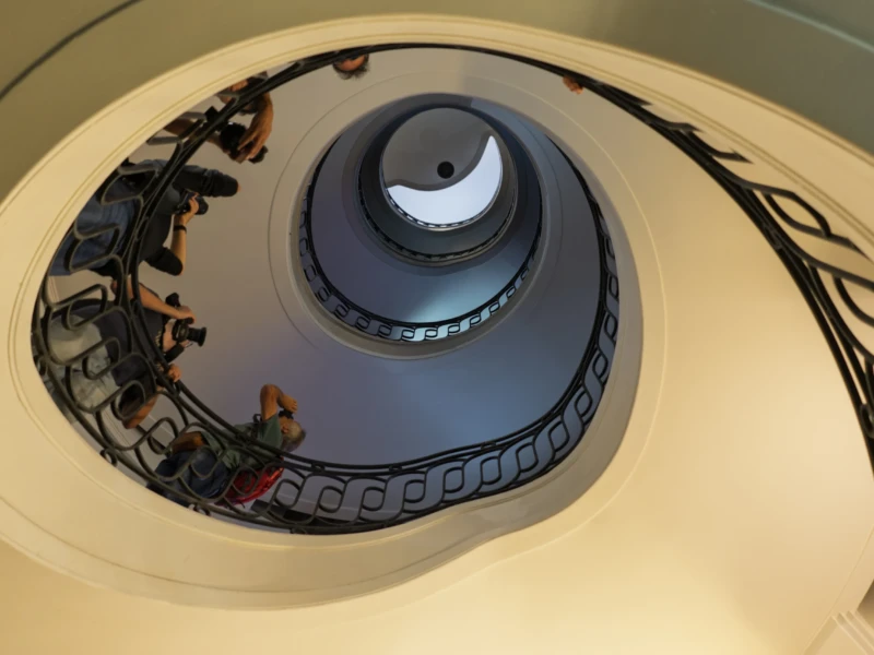 Thierry · 20/09/2026 · 3840 x 2560 Cette photographie montre un escalier hélicoïdal, sujet principal, avec une vue plongeante depuis le haut. La composition met en valeur la symétrie et la profondeur de la structure.Le cadrage est serré, centré sur l’escalier et ses occupants. La lumière est douce et naturelle, éclairant uniformément l’espace. Les couleurs dominantes sont des tons neutres : blanc crème pour les murs, gris pour les marches et balustrades.Le style est épuré et architectural. L’ambiance est à la fois élégante et mystérieuse, renforcée par la présence discrète des personnes.