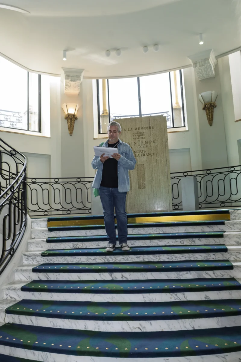 Un homme debout sur un escalier en marbre tenant un document, devant une plaque commémorative dans un bâtiment élégant.