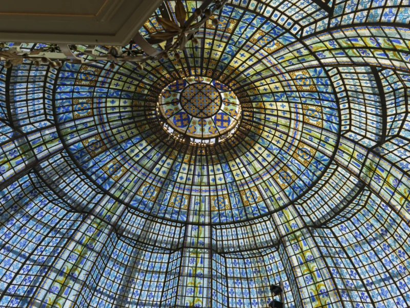 Thierry · 20/09/2026 · 3840 x 2560 Cette photographie montre une vue plongeante vers le haut d’une structure architecturale en forme de spirale. Le sujet principal est une coupole vitrée aux motifs géométriques complexes, composée de nombreux petits panneaux colorés.La composition est centrée et symétrique, avec des lignes convergentes créant une impression de profondeur. Le cadrage est serré, mettant en valeur les détails des vitraux. La lumière, naturelle et filtrée, éclaire uniformément la structure, accentuant les teintes bleutées et dorées. L’ambiance est à la fois géométrique et poétique, évoquant une harmonie entre art et architecture.