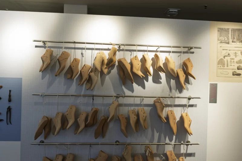 Collection de derniers de chaussures en bois suspendus sur des étagères dans une exposition muséale, éclairés par