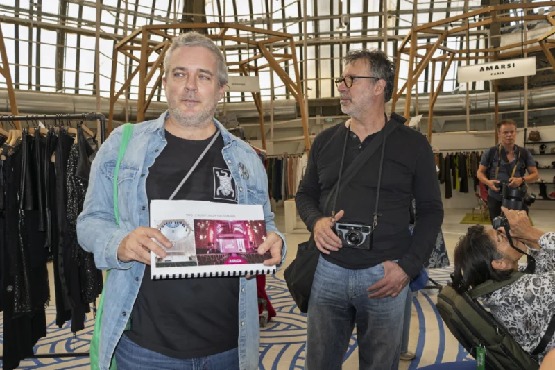 Deux hommes dans un espace d'exposition textile, l'un tenant une affiche et l'autre une vieille caméra, entourés