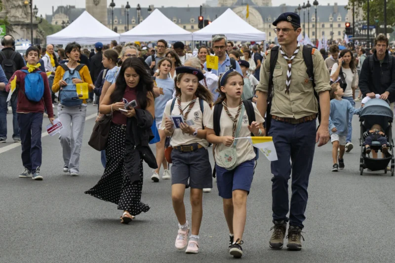 Thierry · 26/09/2026 · 3840 x 2560 Groupe de personnes marchant dans une rue animée, dont des enfants en tenue scout et un adulte en uniforme, entourés