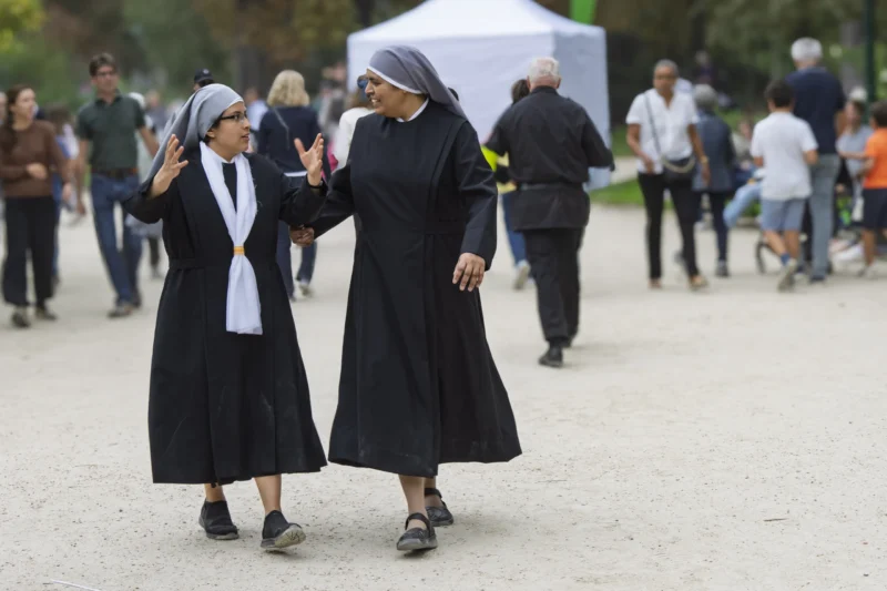 Thierry · 26/09/2026 · 3840 x 2560 Deux religieuses en habit noir et coiffe blanche se tenant par les mains et souriant en extérieur, entourées de passants.
