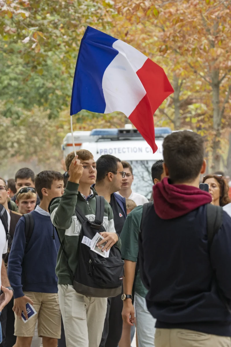 Thierry · 26/09/2026 · 2560 x 3840 Un groupe de personnes en extérieur, dont une tient un drapeau français tricolore levé, dans une ambiance de rassemblement