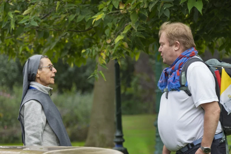 Thierry · 26/09/2026 · 3840 x 2560 Deux personnes engagées dans une conversation sous un arbre dans un cadre verdoyant, l’une vêtue d’un habit religieux